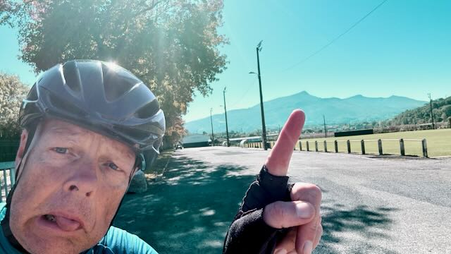 Auf dem Bild ist ein Radfahrer mit Helm und blauer Kleidung zu sehen, der am Straßenrand steht. Er zeigt mit dem Finger auf einen Berg im Hintergrund, der sich deutlich gegen den blauen Himmel abzeichnet. Im Vordergrund verläuft eine leere Straße, daneben einige Bäume und Wiesen, im Hintergrund die Bergkette der Pyrenäen. Das Bild vermittelt den Eindruck eines sonnigen Tages und einer sportlichen Trainingssituation in einer landschaftlich reizvollen Umgebung.